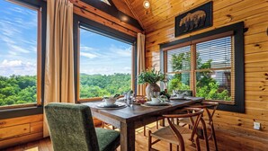 Un intérieur en bois avec une table à manger dressée pour deux personnes, de grandes fenêtres avec des rideaux et une vue sur un paysage boisé.