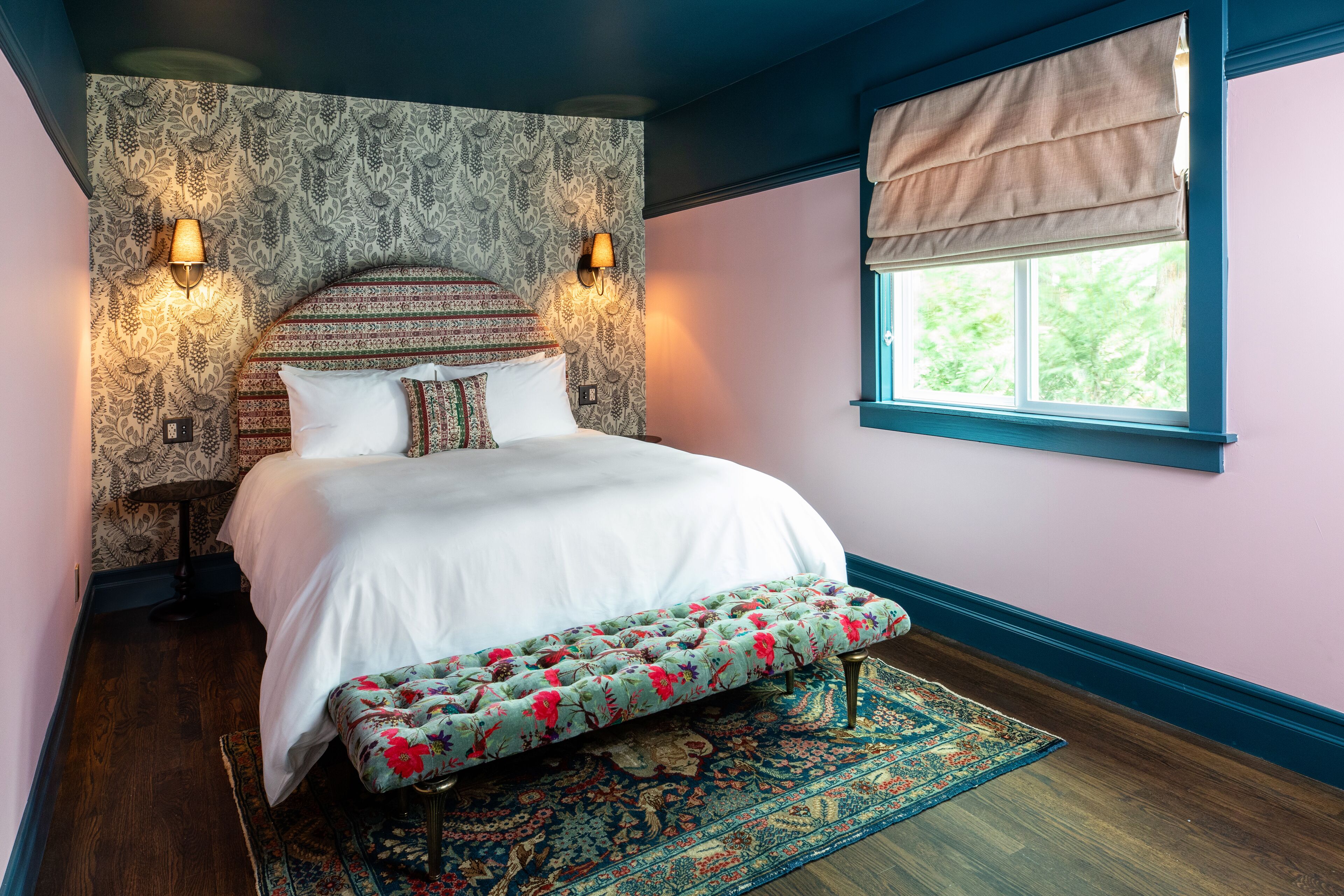 A bedroom with a bed, bench, patterned wallpaper, wall sconces, and a window with a valance.