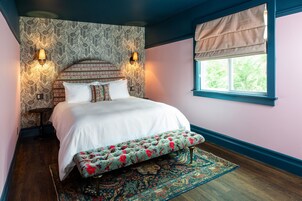 A bedroom with a bed, bench, patterned wallpaper, wall sconces, and a window with a valance.