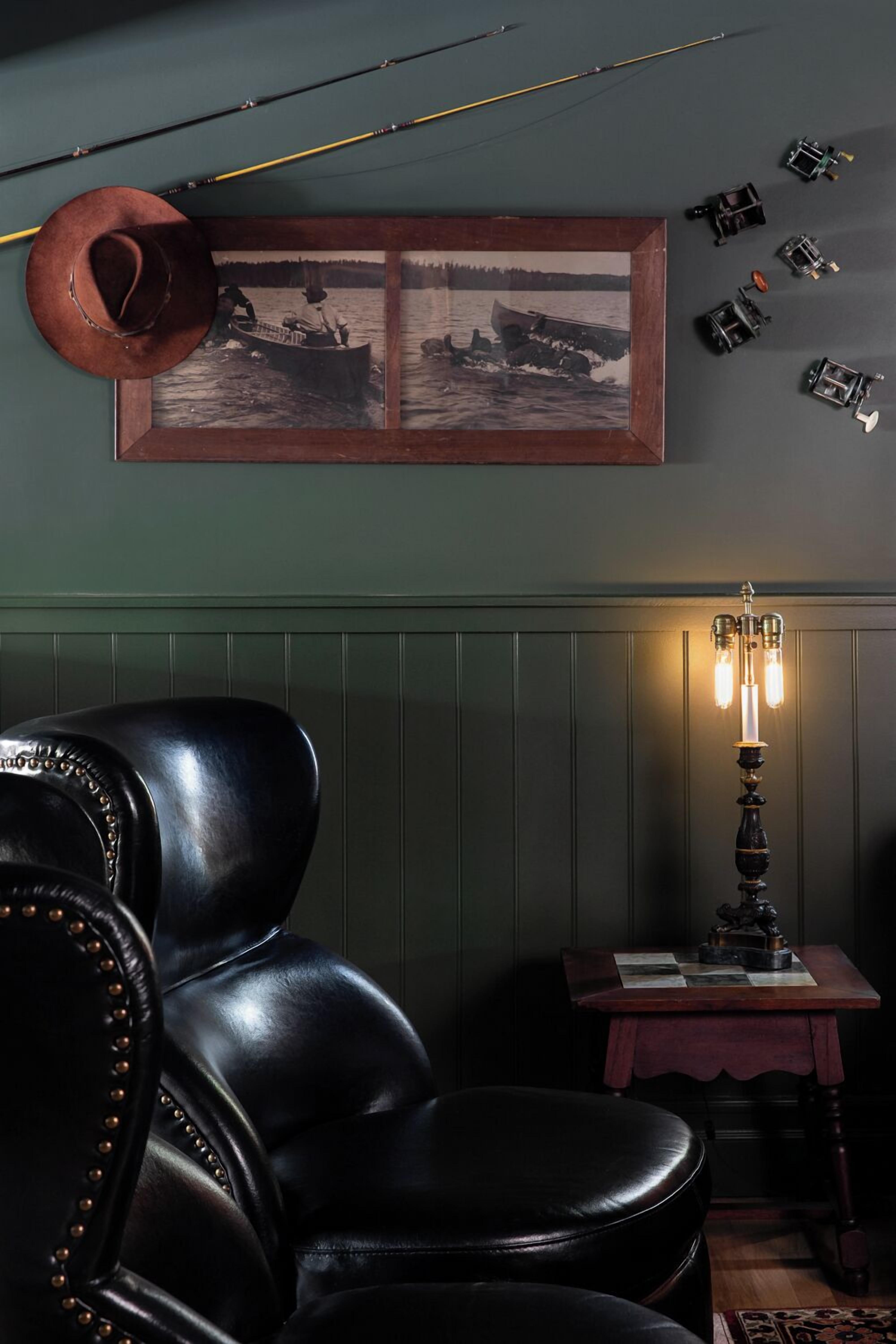 A room with a black leather chair, a wooden table with a lamp, a framed photograph on the wall, and fishing rods.