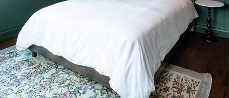 Un dormitorio con papel tapiz estampado con motivos florales, un cabecero verde, una colcha blanca y una mesita redonda con una lámpara.