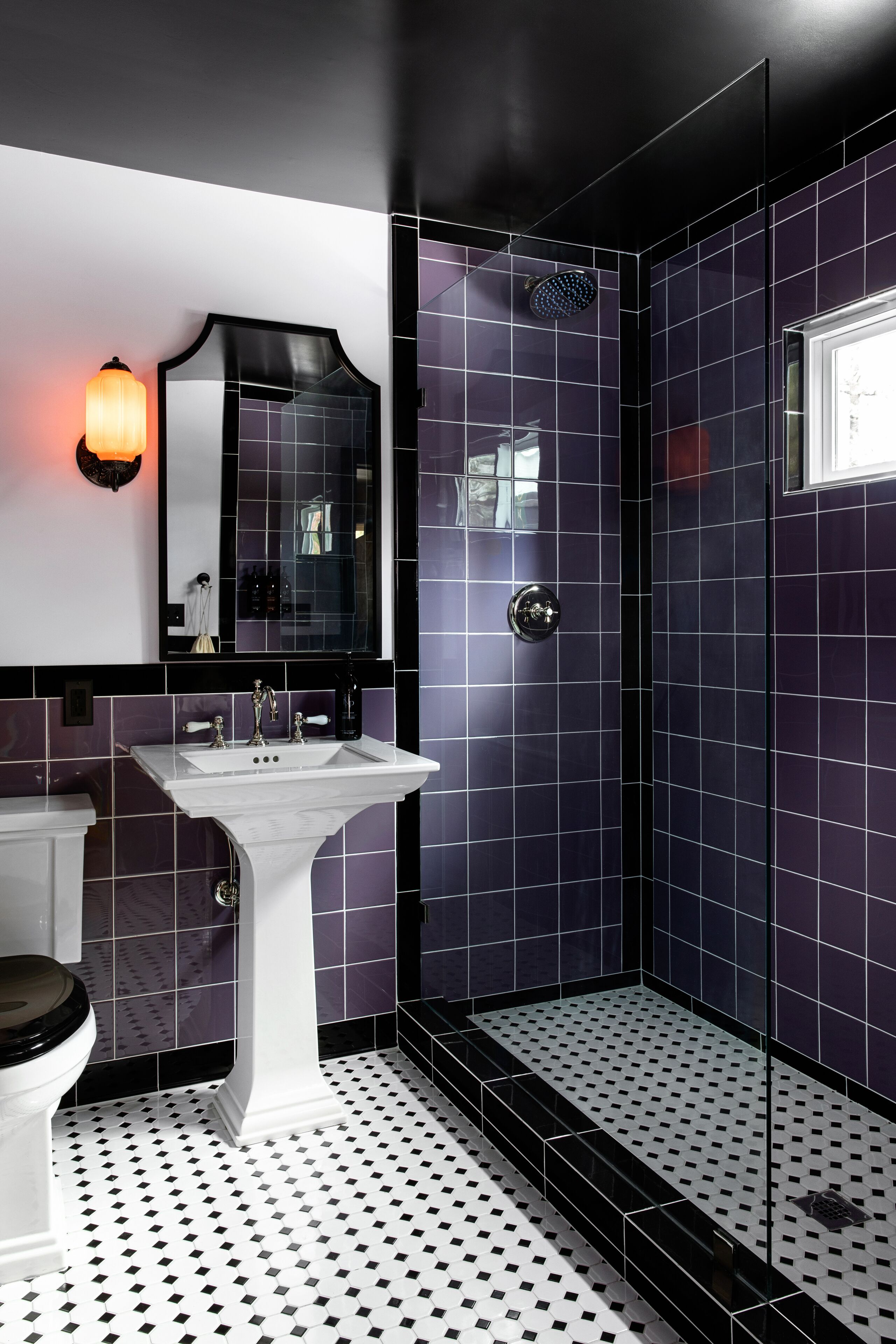 Une salle de bain avec un lavabo sur pied blanc, des toilettes noires et une douche séparée par des parois en verre, dont les murs sont recouverts de carreaux violets.
