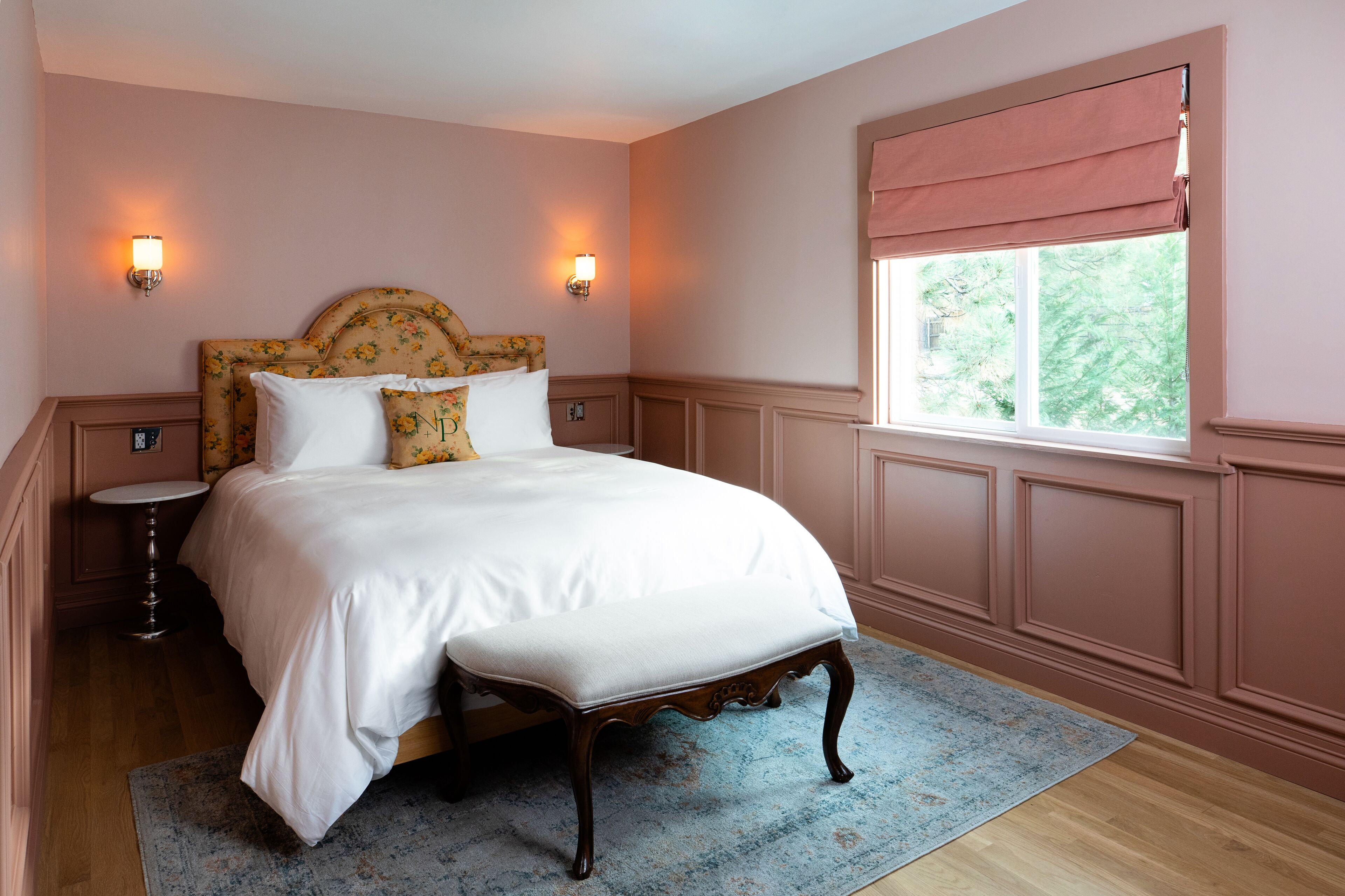 A bedroom with a large bed, two bedside tables, a window with a view, and wall sconces.