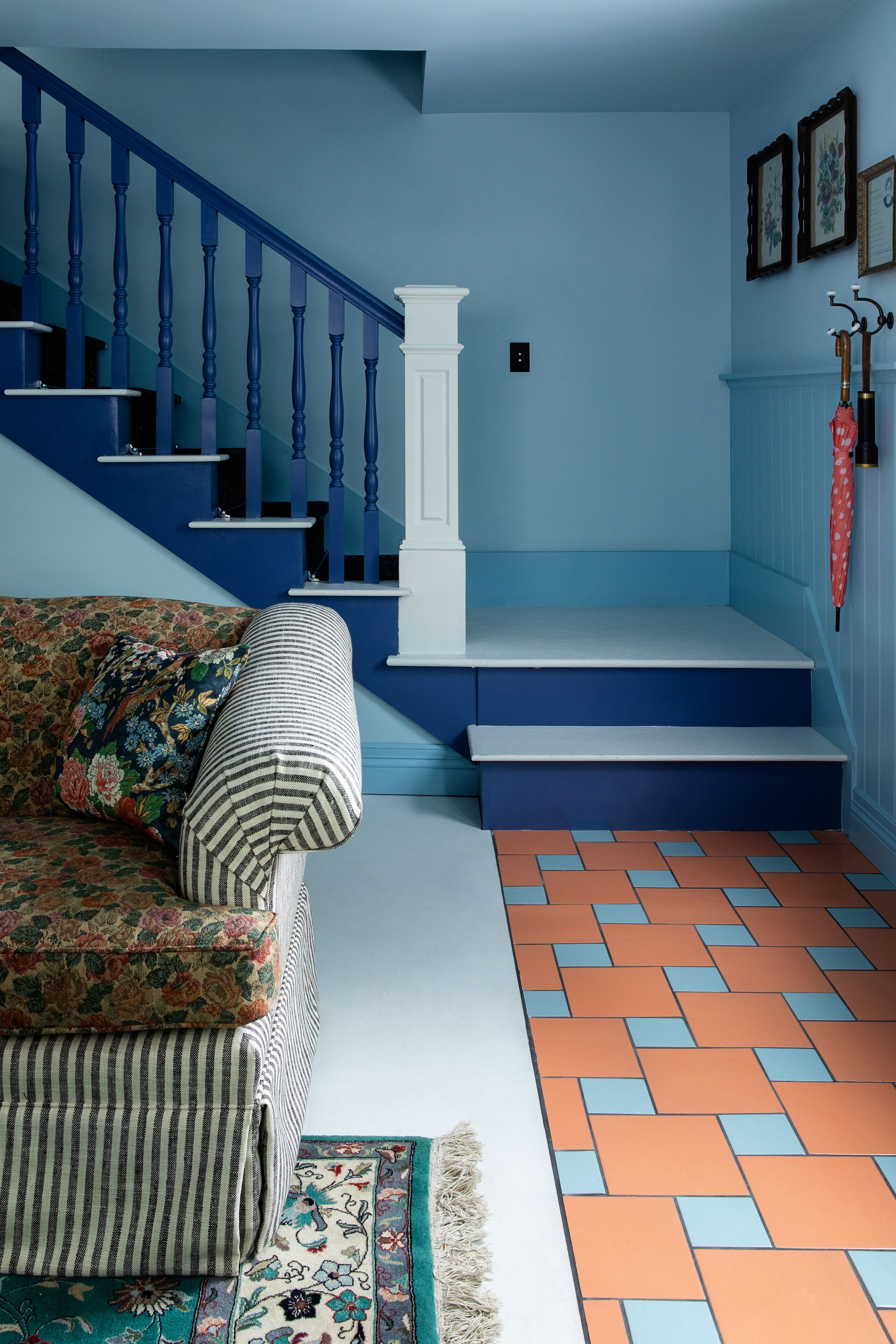 A living room with a blue staircase, a floral sofa, a striped armchair, and a patterned rug.