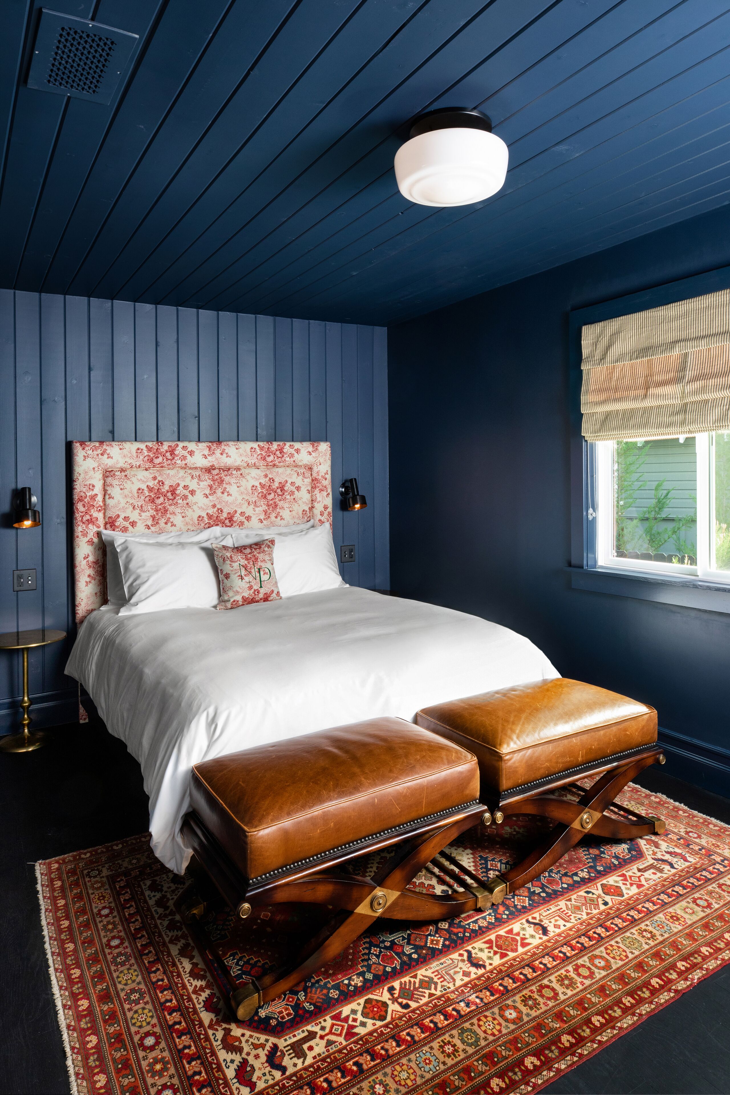 A bedroom with a large bed, two brown leather ottomans, a patterned rug, and a window with a view.