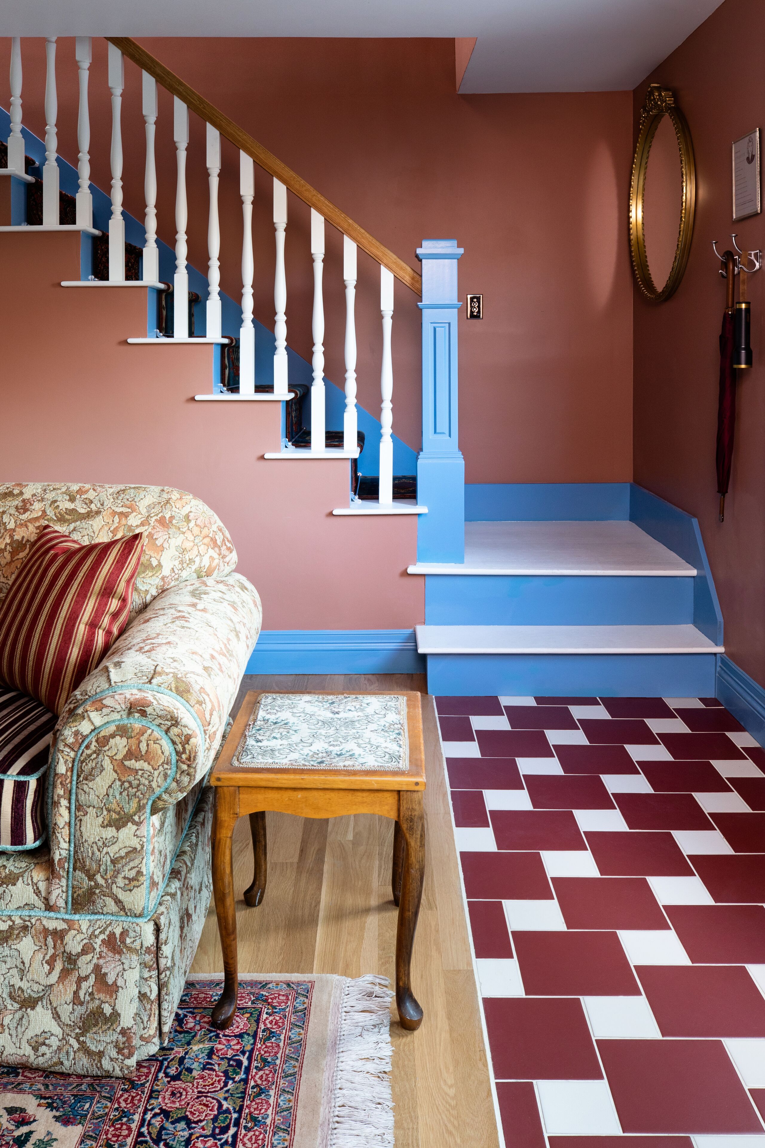 A staircase with white balusters and blue steps, a floral sofa with a patterned rug, and a herringbone-patterned floor.