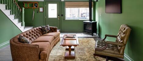 Una sala de estar con un sofá de cuero marrón, una mesa de centro de madera y un sillón de cuero. Se ve una escalera con paredes verdes y barandilla de madera.