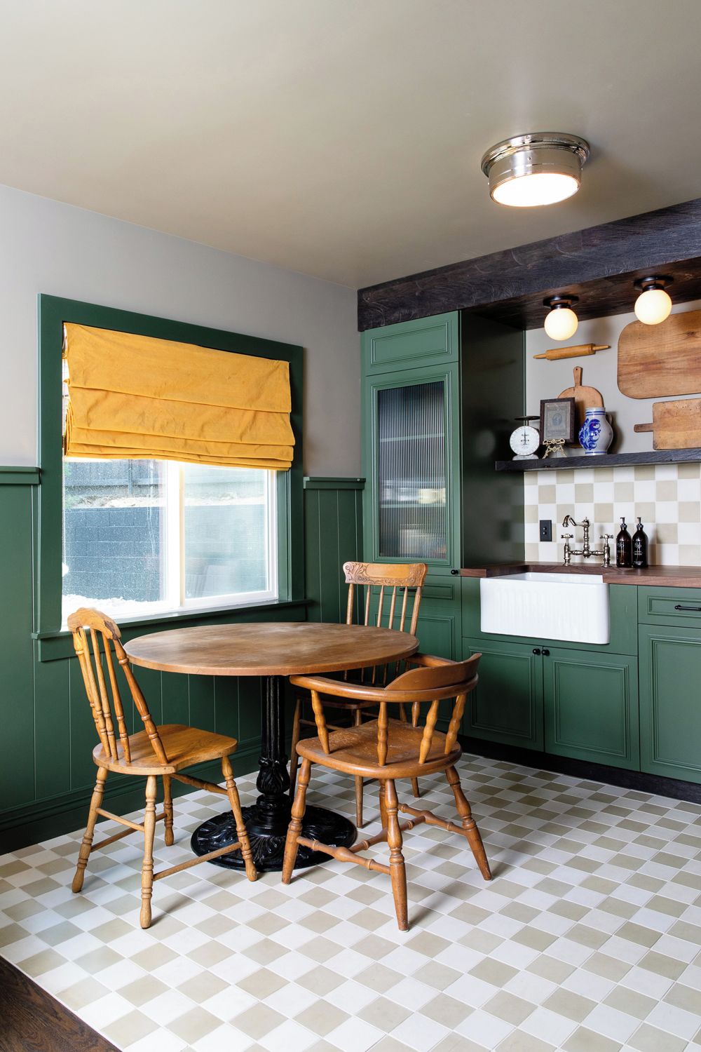 A kitchen with a round table and four wooden chairs, a window with yellow curtains, green cabinets, and a checkered floor.