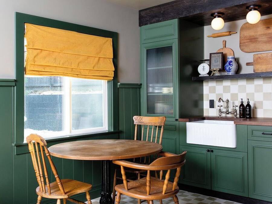 A kitchen with a round table and four wooden chairs, a window with yellow curtains, green cabinets, and a checkered floor.