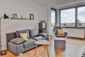 Relax in the living room with a sofa, armchair, and floor lamp, surrounded by a large window with a view of the sky and distant landscape.
