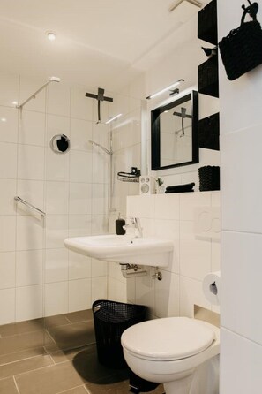 A modern bathroom with a white sink, toilet, and black trash bin, set against a backdrop of white-tiled walls and beige flooring.