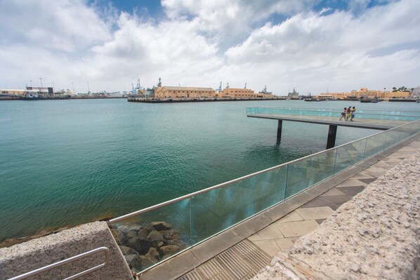 A waterfront promenade with a glass railing overlooking calm water, a stone pathway, and distant buildings. - Cian Beach Home By CanariasGetaway (Las Palmas de Gran Canaria)