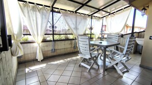 En udendørs terrasse med et bord og stole, et vindue med gardiner, og et flisegulv.