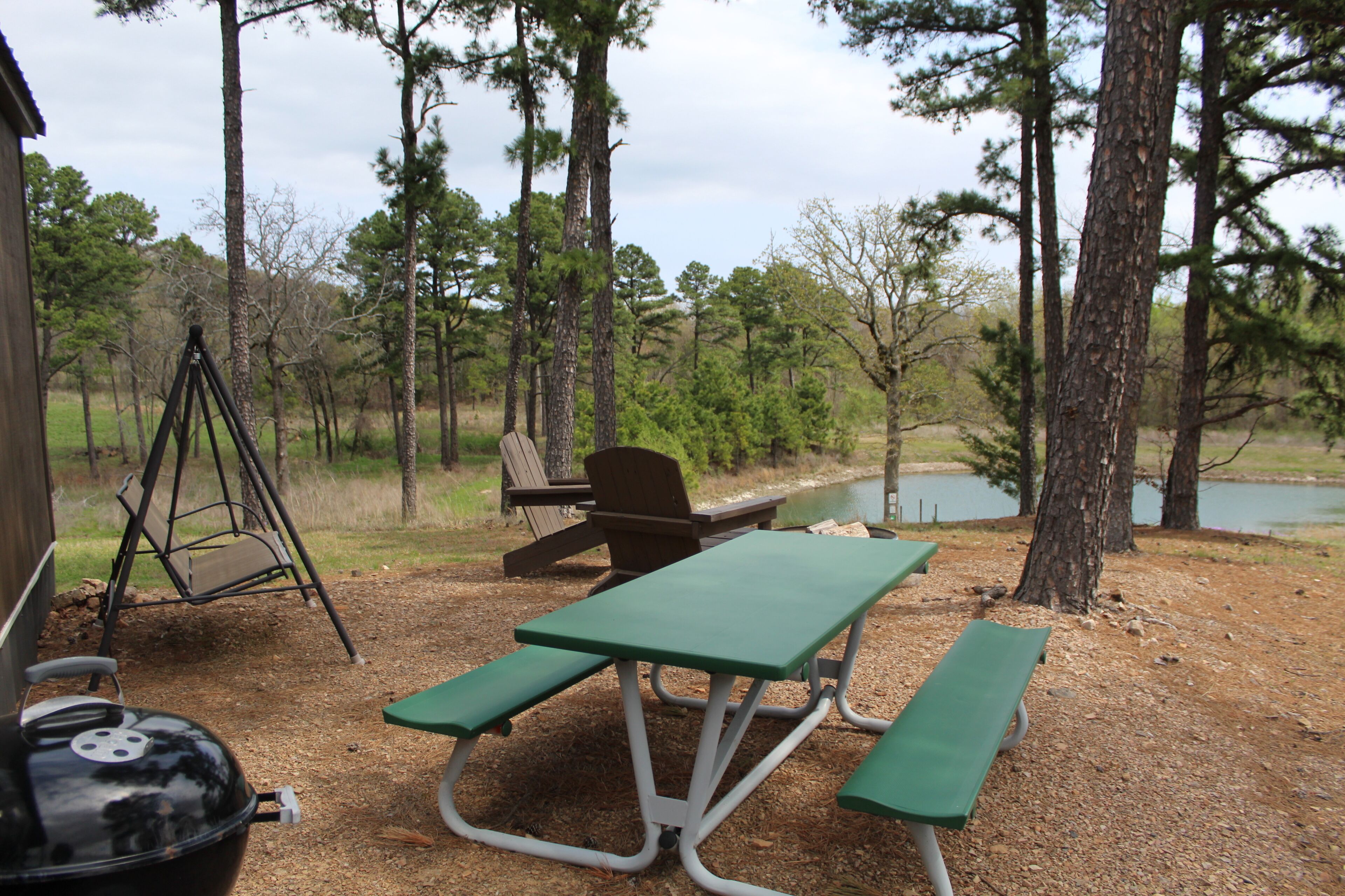 Une table de pique-nique avec des bancs, une balançoire et un barbecue, le tout installé dans un espace boisé près d’un lac.