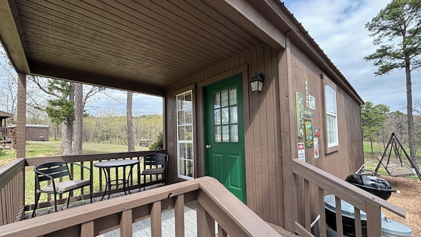 A wooden cabin with a green door, a covered porch with a table and chairs, and a view of the surrounding landscape. - Fish Cabin at Mountain View Retreat (Talihina)