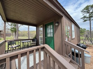 A wooden cabin with a green door, a covered porch with a table and chairs, and a view of the surrounding landscape. - Fish Cabin at Mountain View Retreat (Talihina)