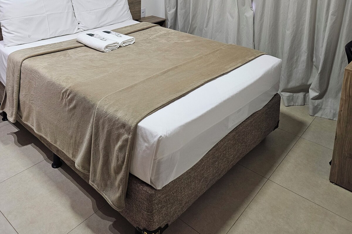 Una cama bien hecha con una manta color beige y almohadas blancas, un cabecero de madera y una ventana con cortinas de colores claros.
