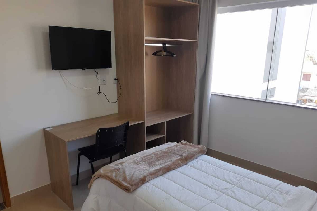 Un dormitorio con cama, escritorio, silla, televisor montado en la pared y ventana.