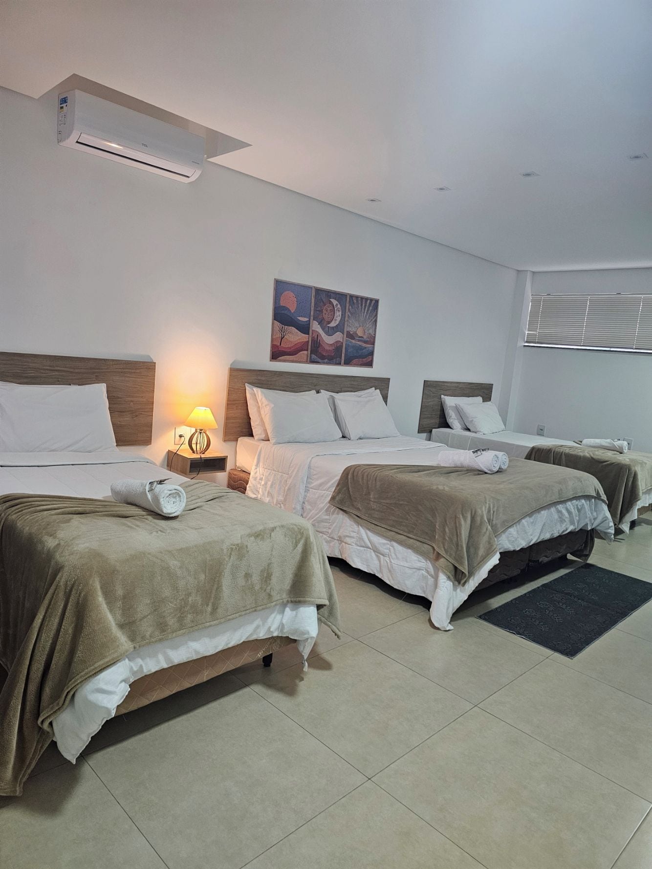 A hotel room with three beds, a wall-mounted air conditioner, a nightstand with a lamp, and a painting above the beds.