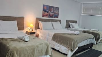 A hotel room with three beds, a wall-mounted air conditioner, a nightstand with a lamp, and a painting above the beds.