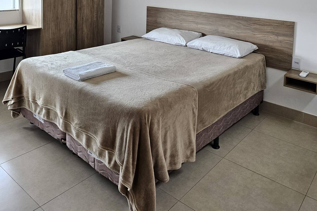 Una cama bien hecha con cabecera de madera, una mesita de noche con un teléfono y un aire acondicionado fijado a la pared.