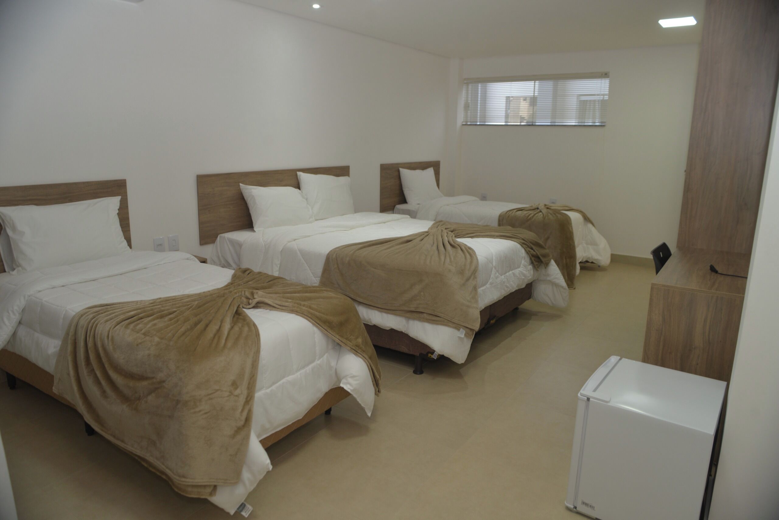 Habitación de hotel con tres camas, un refrigerador pequeño y una ventana con persianas.