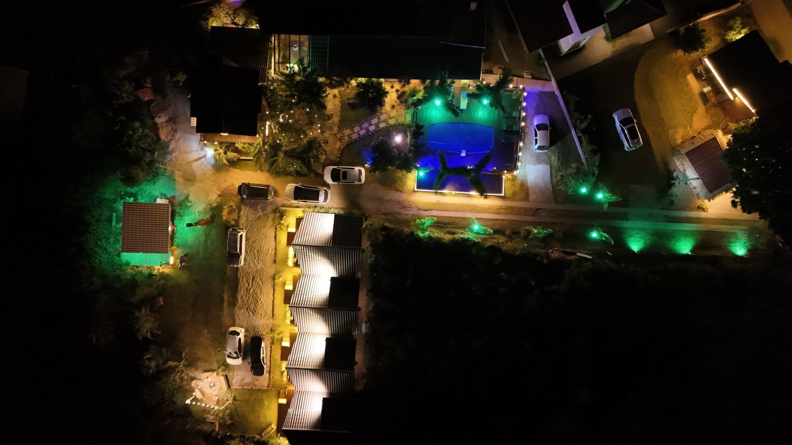 An aerial view of a residential area with a swimming pool, multiple cars parked, and a house with a green roof.