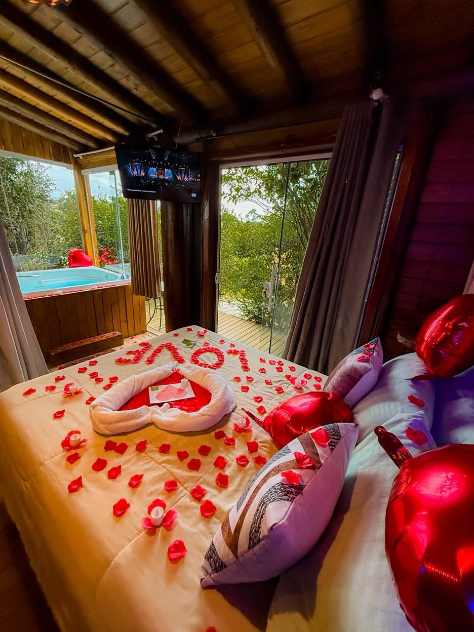 Ein gemütliches Schlafzimmer mit einem Bett, das mit Rosenblüten und einem herzförmigen Kissen dekoriert ist, einem Fernseher an der Wand und Blick auf einen Pool im Freien.