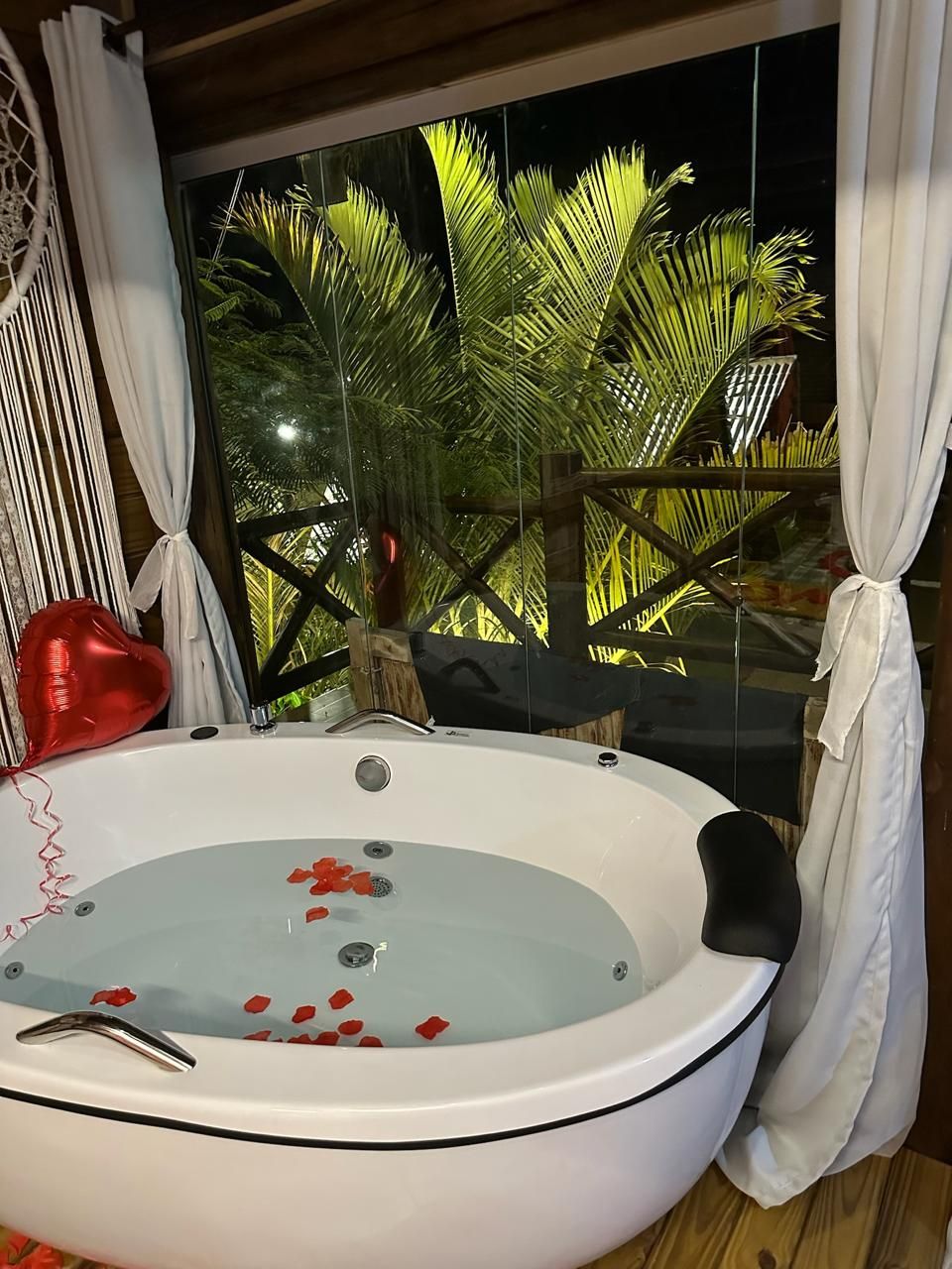Un jacuzzi con pétalos de rosa, rodeado de cortinas blancas y vistas a palmeras.