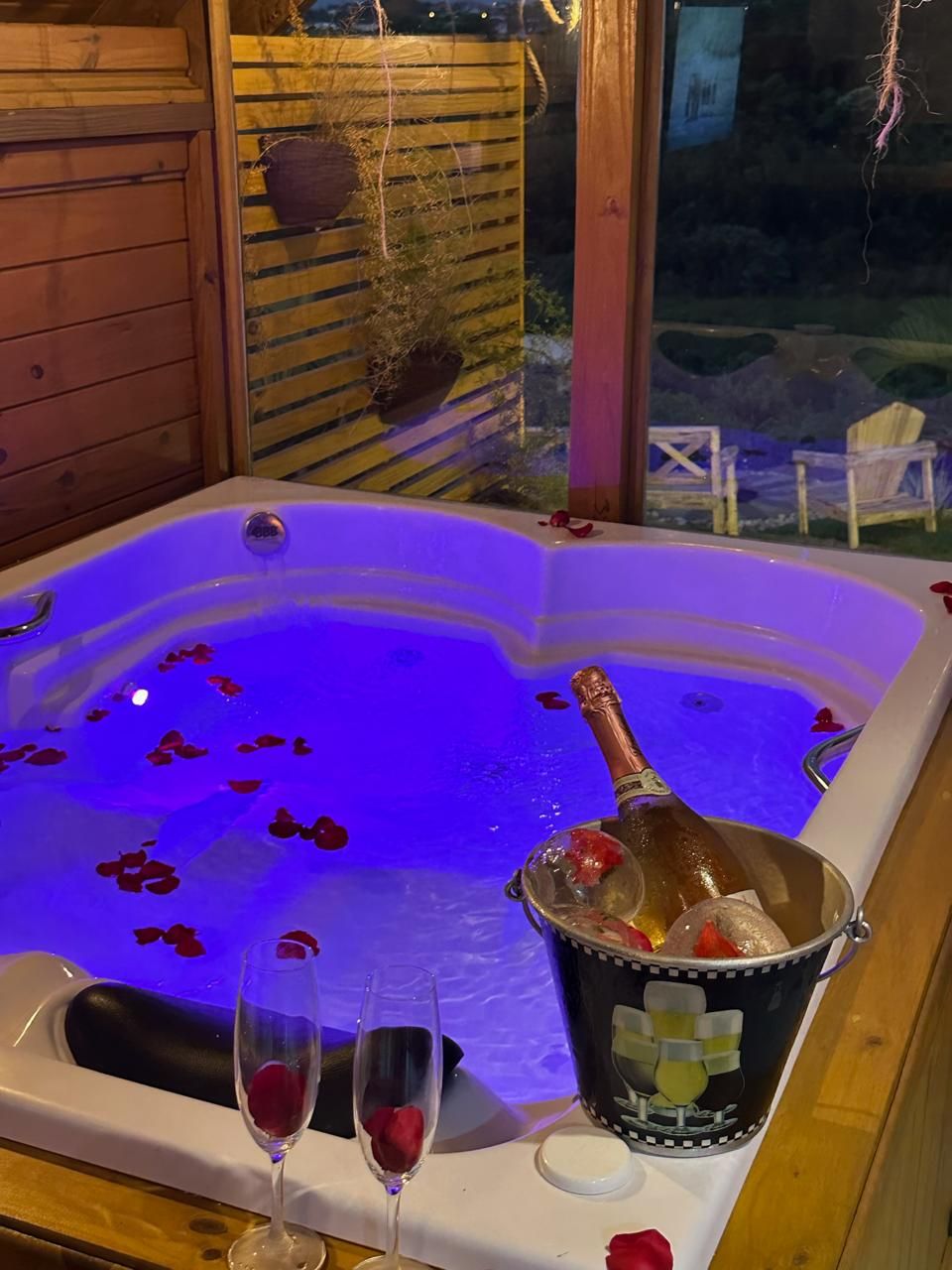 A hot tub with rose petals and champagne, surrounded by wooden walls and a view of the outdoors.