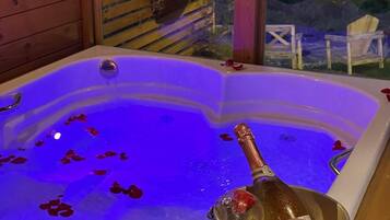 A hot tub with rose petals and champagne, surrounded by wooden walls and a view of the outdoors.