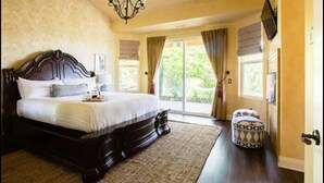 A bedroom with a large bed, a chandelier, a window with curtains, and a door with a television. - Italian Estate in Wine Country (Temecula)