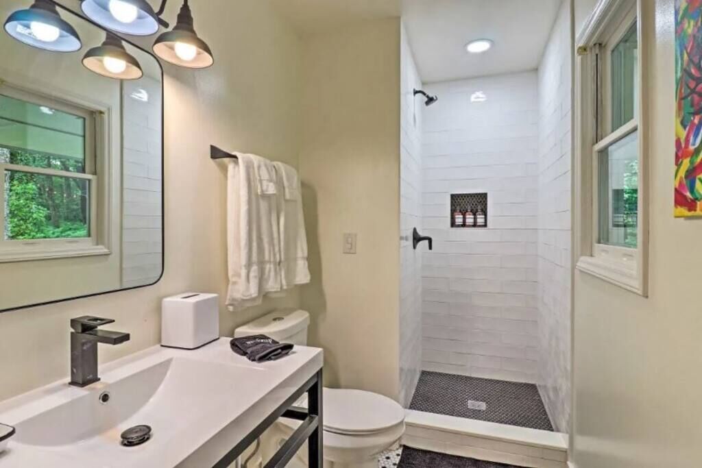 A bathroom with a white sink, a toilet, and a shower. There are three lights above the sink and a window on the left side.