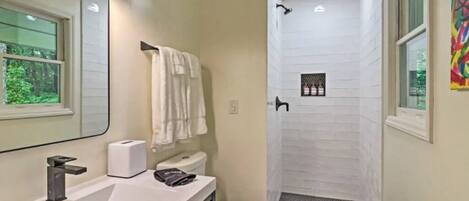 A bathroom with a white sink, a toilet, and a shower. There are three lights above the sink and a window on the left side.