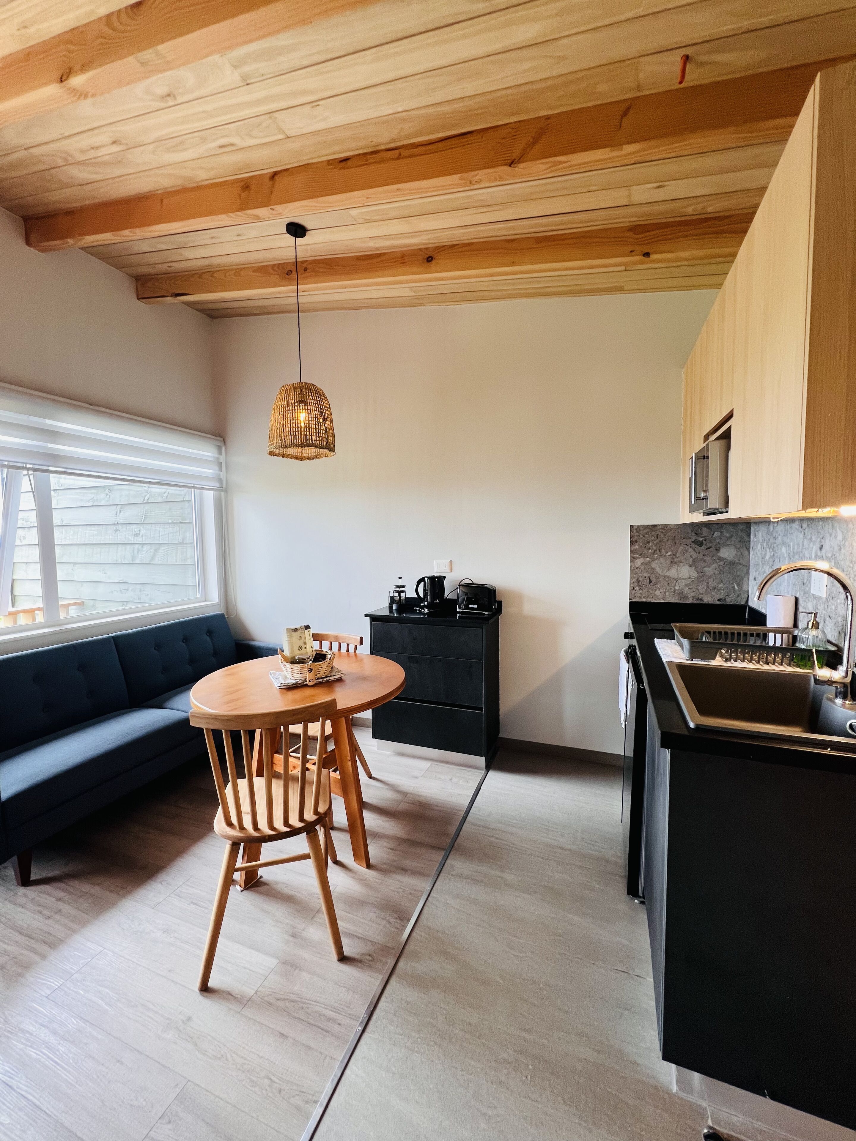 Une cuisine moderne avec un plafond en bois, un coin repas avec une table ronde et des chaises, un meuble noir et une fenêtre avec des stores.