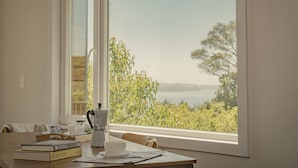 Una mesa con libros, una cafetera y una taza, cerca de una ventana con vistas a árboles y una masa de agua.