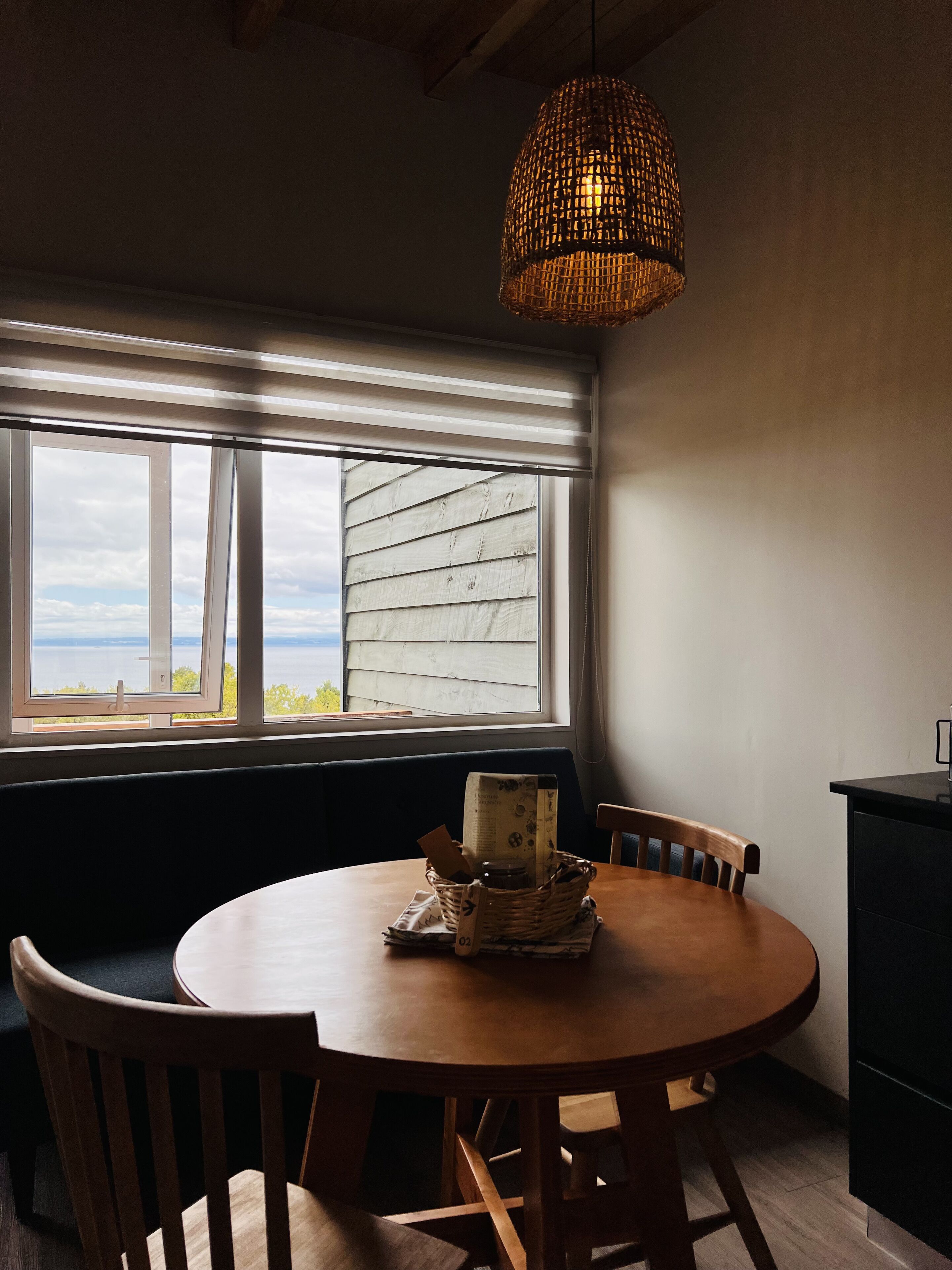 Un comedor con una mesa redonda de madera, una lámpara colgante de fibra y vistas a través de una ventana con persianas.