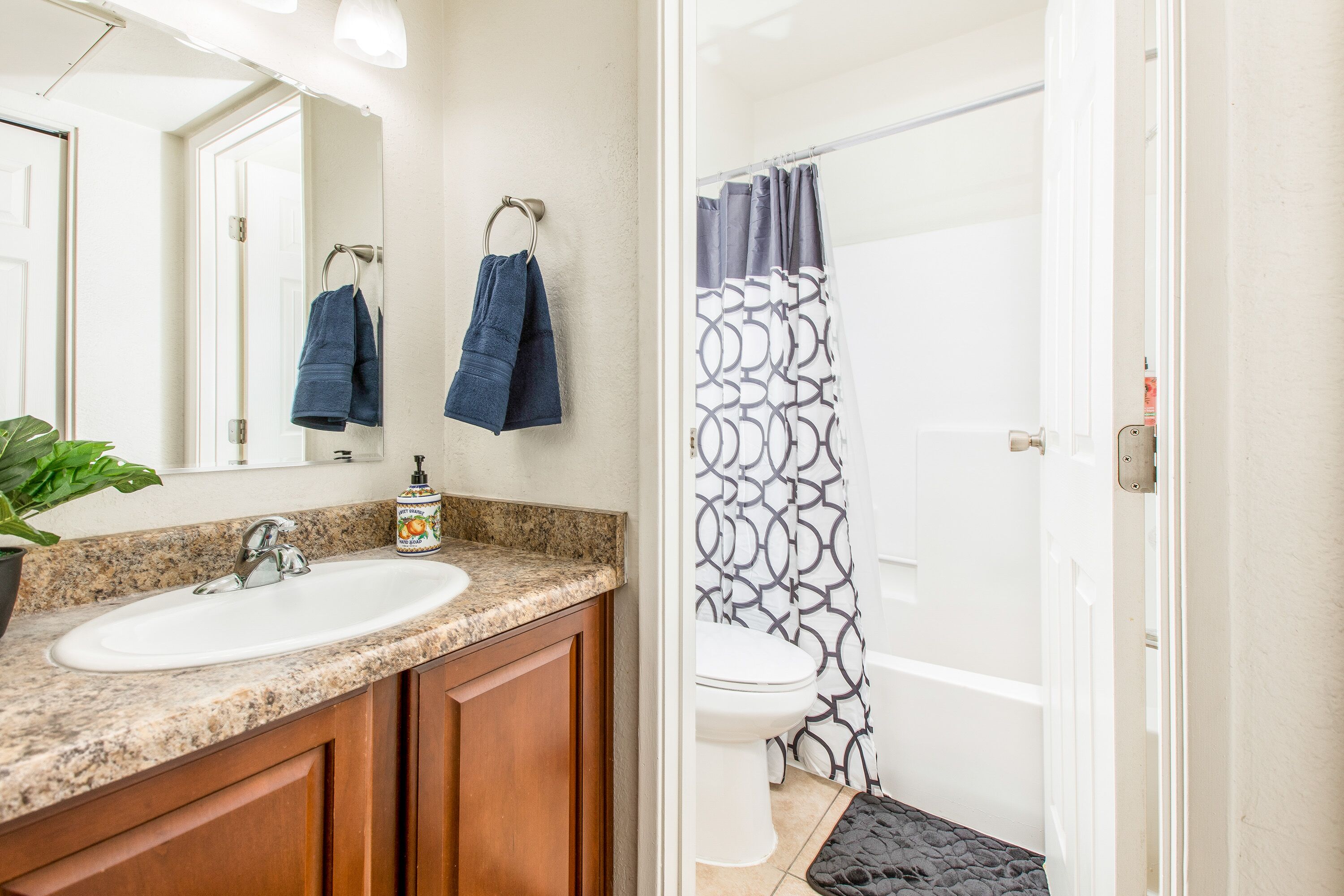 Une salle de bain avec un lavabo blanc, des meubles en bois, un miroir, des toilettes et une douche avec un rideau à motifs.