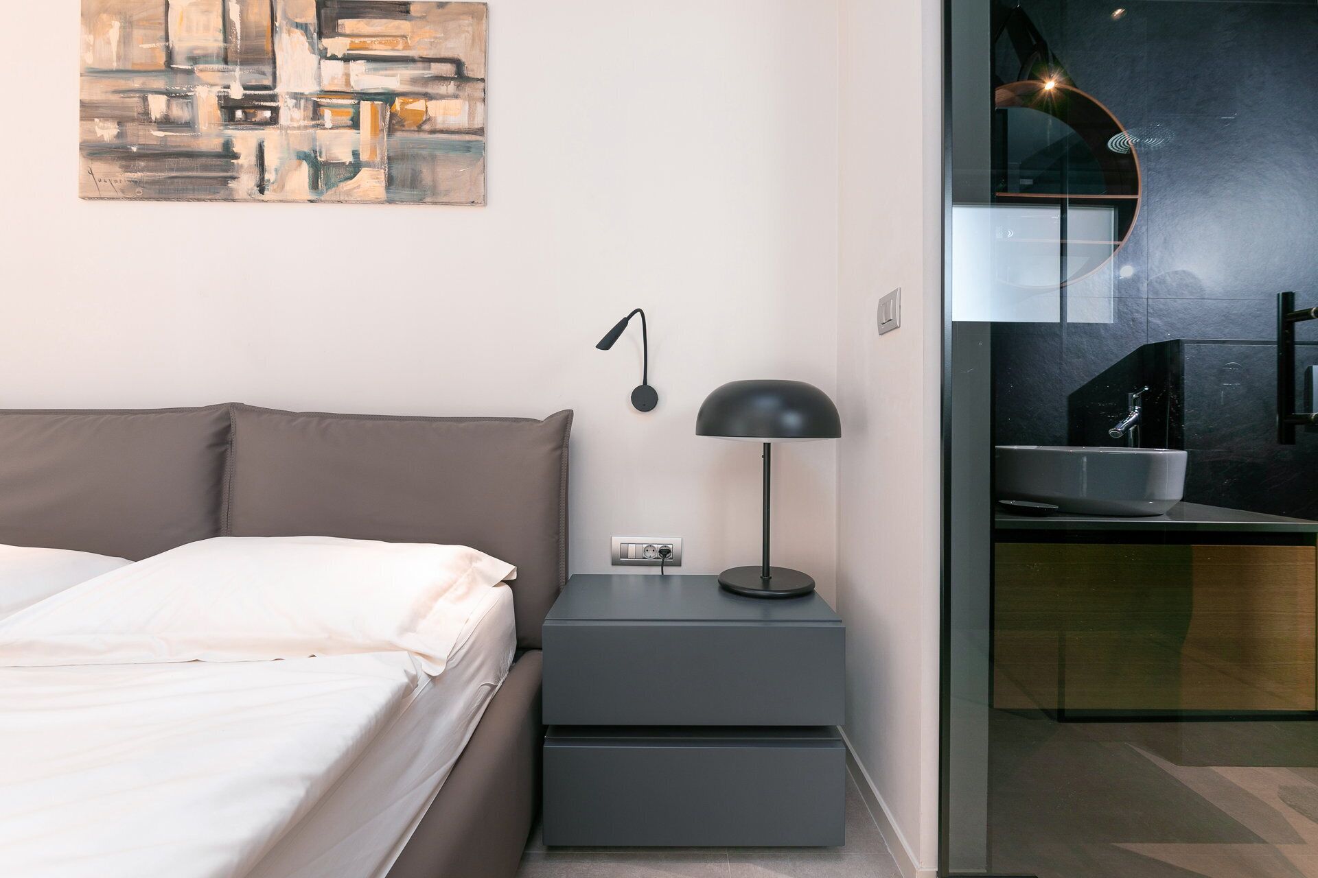 Un dormitorio moderno con cama, mesita de noche, lámpara y vistas al baño.