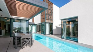 Un área de piscina moderna al aire libre, con mobiliario para comidas y habitaciones con paredes de vidrio.