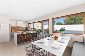 A modern kitchen with a central island, dining area with a long table set for four, and large windows offering a view of the outdoors.