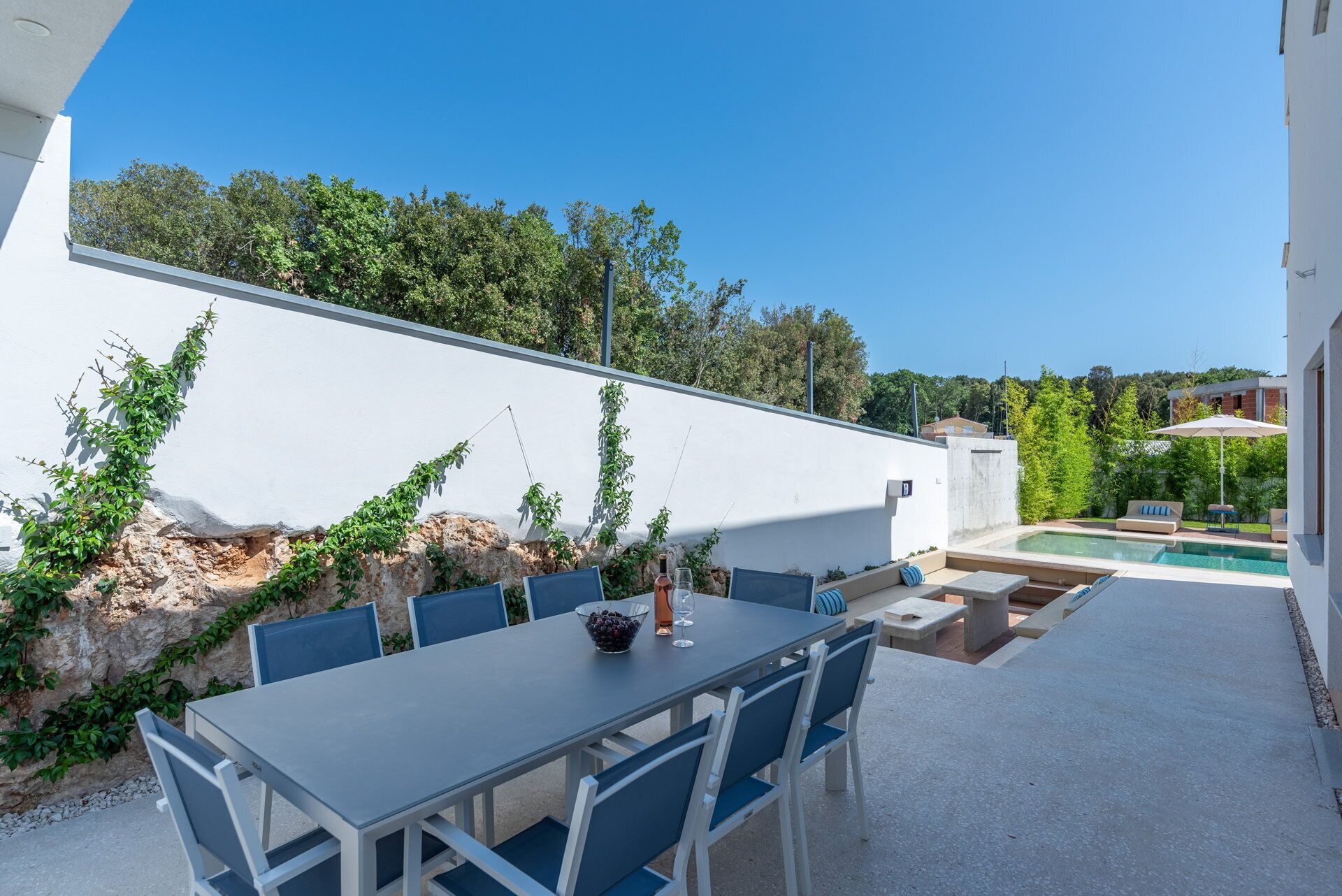 Um pátio externo com mesa e cadeiras de jantar, piscina e uma parede branca com vegetação.