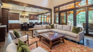 A spacious living area with a large sectional sofa, a coffee table, and a fireplace. There is a kitchen area with dark wooden cabinets and a large window overlooking a forested area.