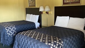 A hotel room with two beds, each with a patterned bedspread and white pillows. There is a nightstand with a lamp between the beds. The room has yellow walls and a ceiling with a grid pattern. - BUDGETINN (Monroeville)