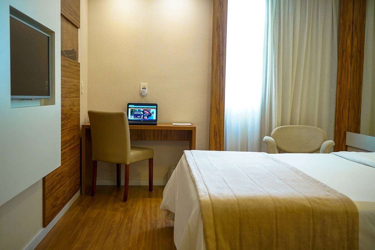 Een hotelkamer met een bed, een bureau met een laptop, een stoel en een raam met gordijnen.