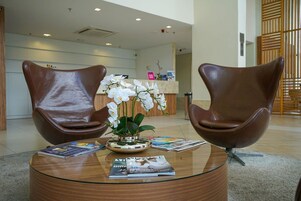 Uma área de recepção com duas poltronas de couro marrom, uma mesa de centro de vidro com revistas, e um vaso de orquídeas brancas.