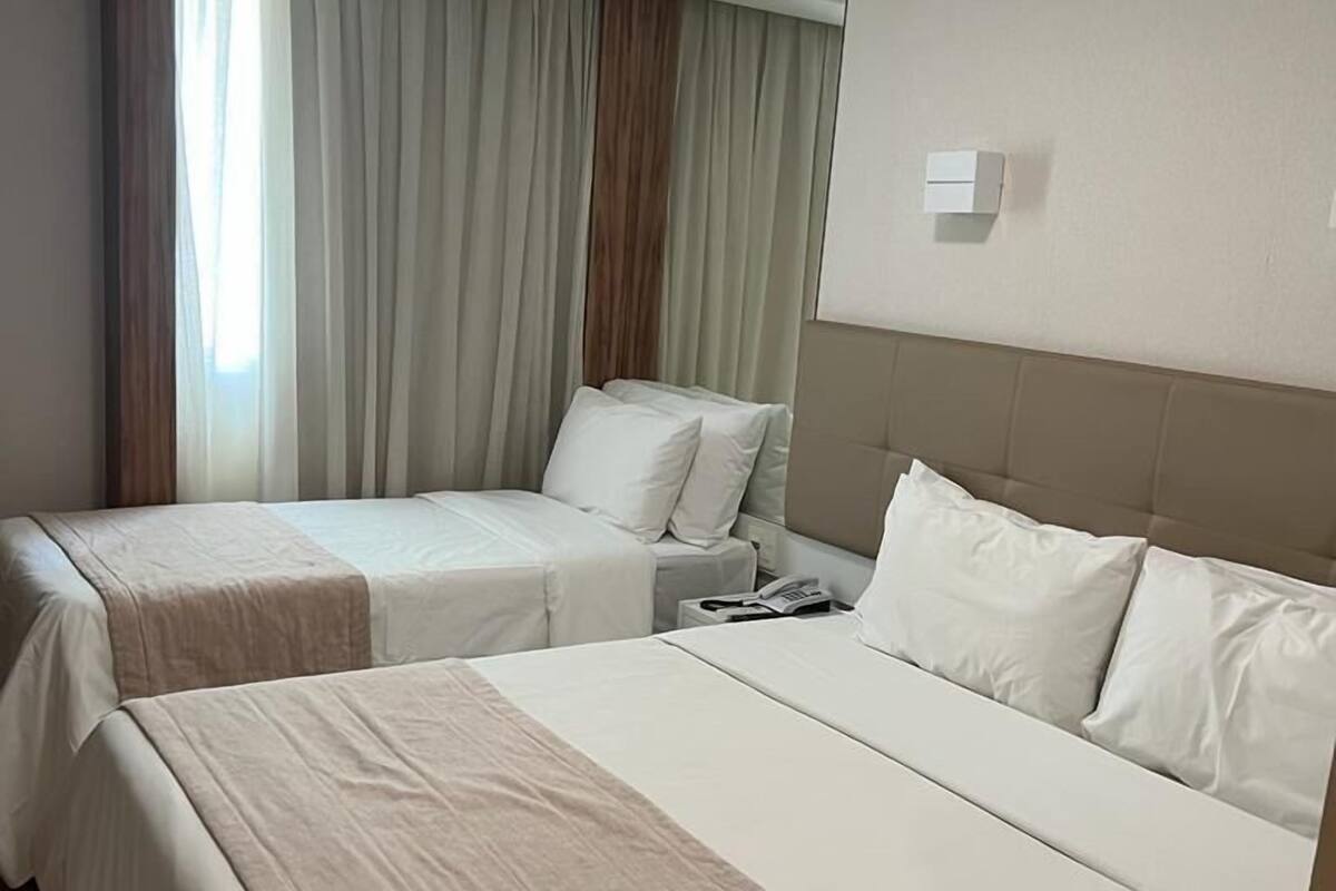 A hotel room with two beds, a large window with curtains, a bedside table with a lamp, and a wall-mounted light fixture.