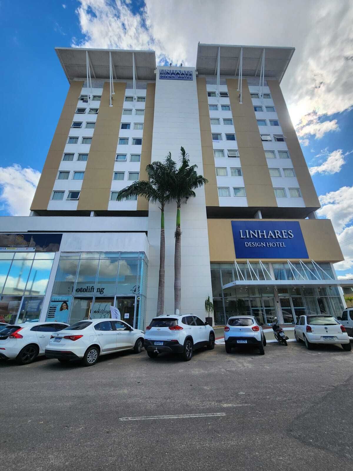 A tall hotel building with a sign reading "LINHARES DESIGN HOTEL". There are several cars parked in front, and a palm tree is visible in front of the building.