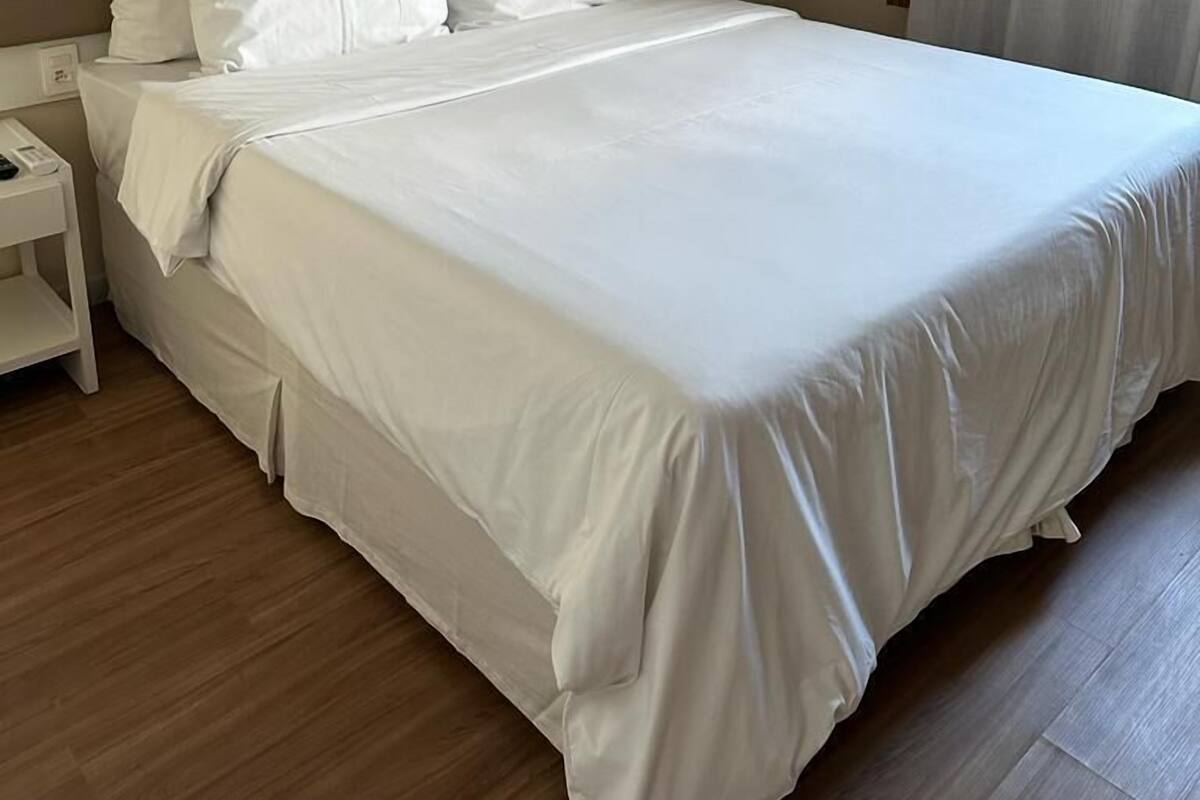 A neatly made bed with white linens in a hotel room with a wooden floor and a beige headboard.