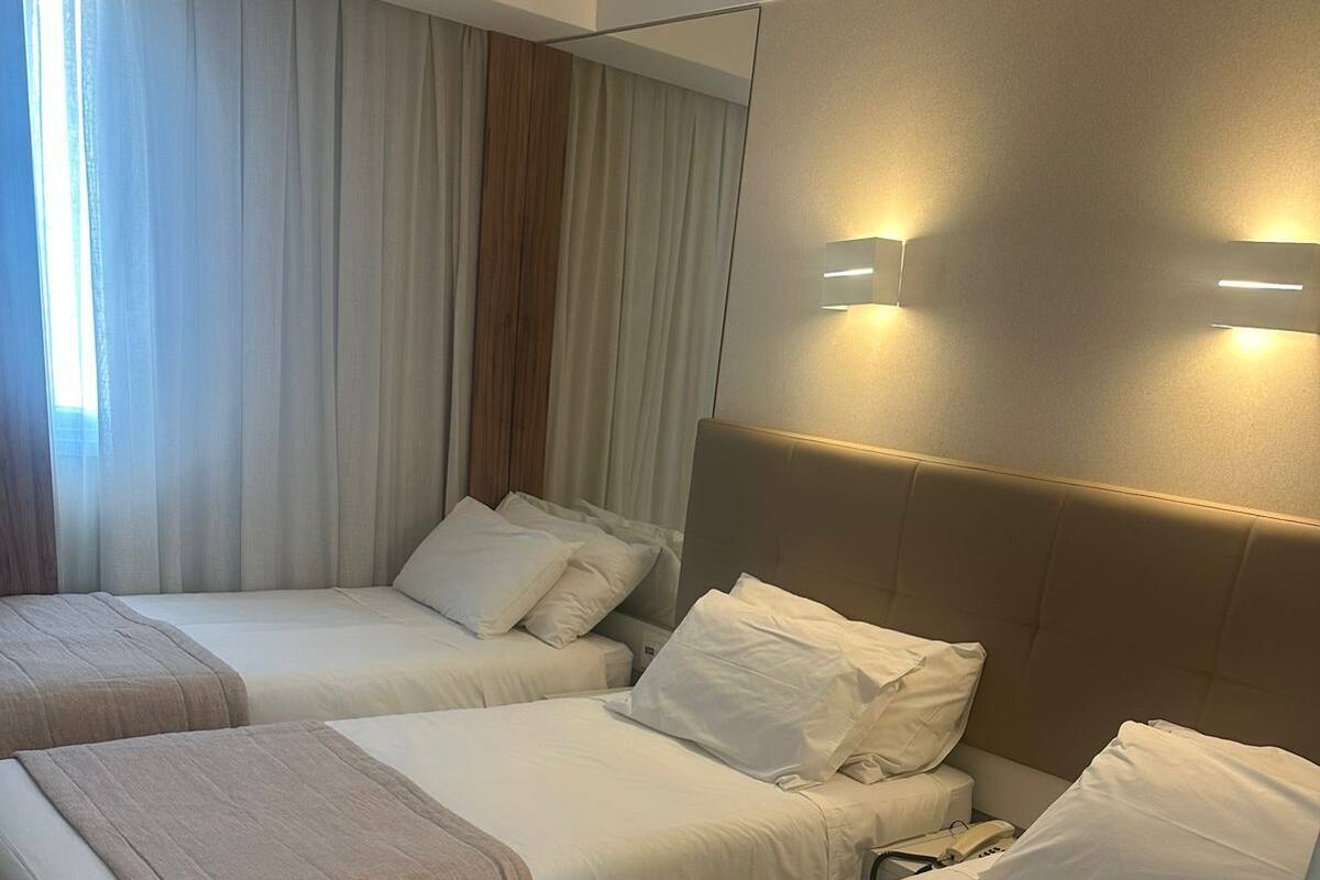 A hotel room with three single beds, each with a headboard and white bedding. There is a large window with curtains, and a wall-mounted light fixture above the beds.